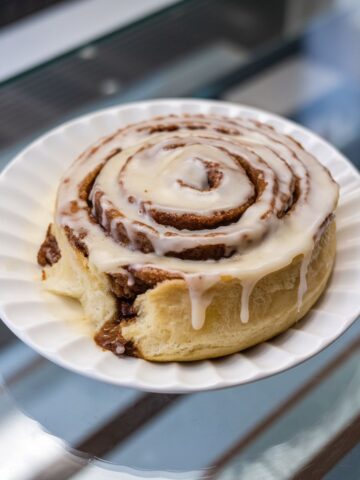 Close-up of a freshly baked cinnamon roll on a white plate, swirled with creamy icing dripping over its soft, golden-brown edges.