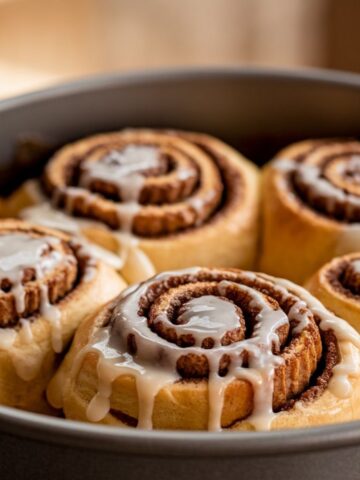 reate a close-up image of freshly baked cinnamon rolls in a round baking pan. The rolls are golden-brown, swirled with cinnamon filling, and generously drizzled with creamy, glossy vanilla icing melting down the sides. The setting is warm and inviting, with soft natural light emphasizing the texture and details of the rolls. The background is blurred to focus attention on the gooey, delicious cinnamon rolls.