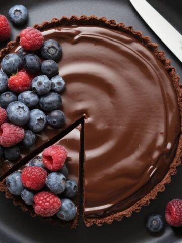 A chocolate tart topped with fresh raspberries and blueberries, with a slice cut out, placed on a black plate with a knife beside it.