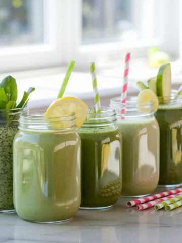 Five mason jars filled with vibrant green smoothies, each topped with unique garnishes like lemon slices, spinach leaves, and cucumber, arranged on a bright kitchen countertop with colorful straws.