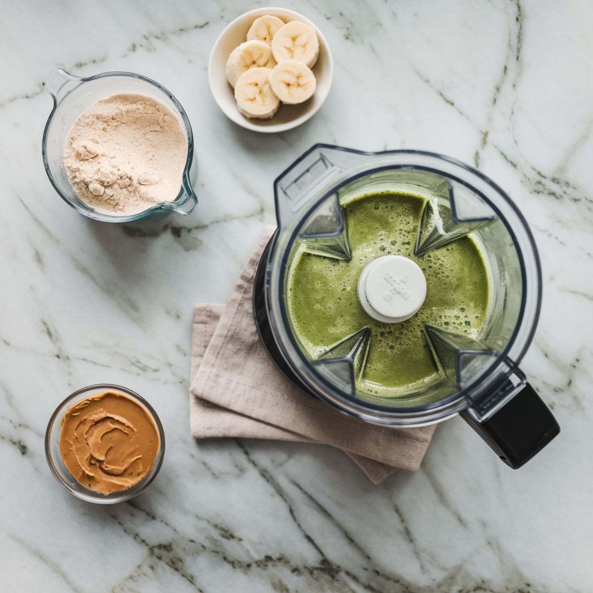 A frozen banana, protein powder, and almond butter added to the blender with the green spinach mixture, creating a creamy texture.