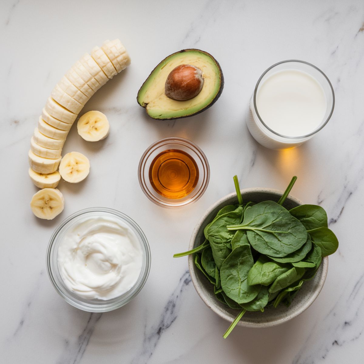 Chopped banana, avocado, and measured yogurt, honey, and almond milk on a marble counter with washed spinach leaves.