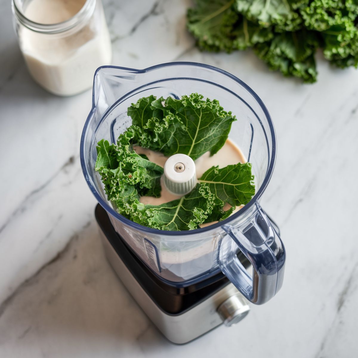 A blender on a marble countertop, filled with chopped kale and coconut milk, with extra kale and a jar of coconut milk in the background.