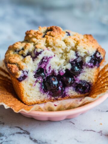 A close-up of a blueberry muffin with a golden streusel topping, torn open to reveal a fluffy, berry-filled center.