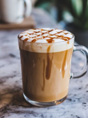 A caramel macchiato in a clear glass mug, topped with frothy milk and caramel drizzle, sitting on a marbel counter.