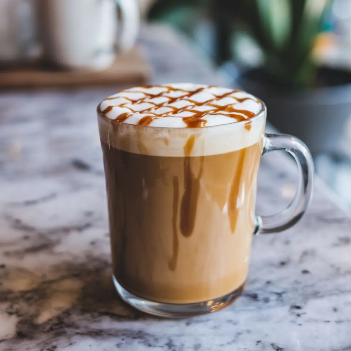  A caramel macchiato in a clear glass mug, topped with frothy milk and caramel drizzle, sitting on a marbel counter.