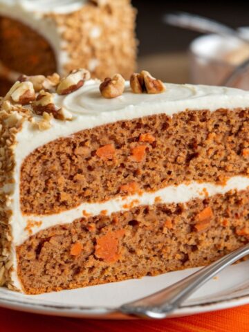 Homemade carrot cake slice with cream cheese frosting, topped with walnuts, on a decorative plate with a fork, set against a warm, inviting background.