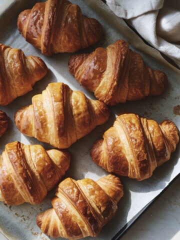 A baking tray lined with parchment paper holds several golden-brown croissants with flaky layers. Soft morning light highlights their crust, and a folded cloth napkin lies beside the tray, evoking a warm, inviting feel.