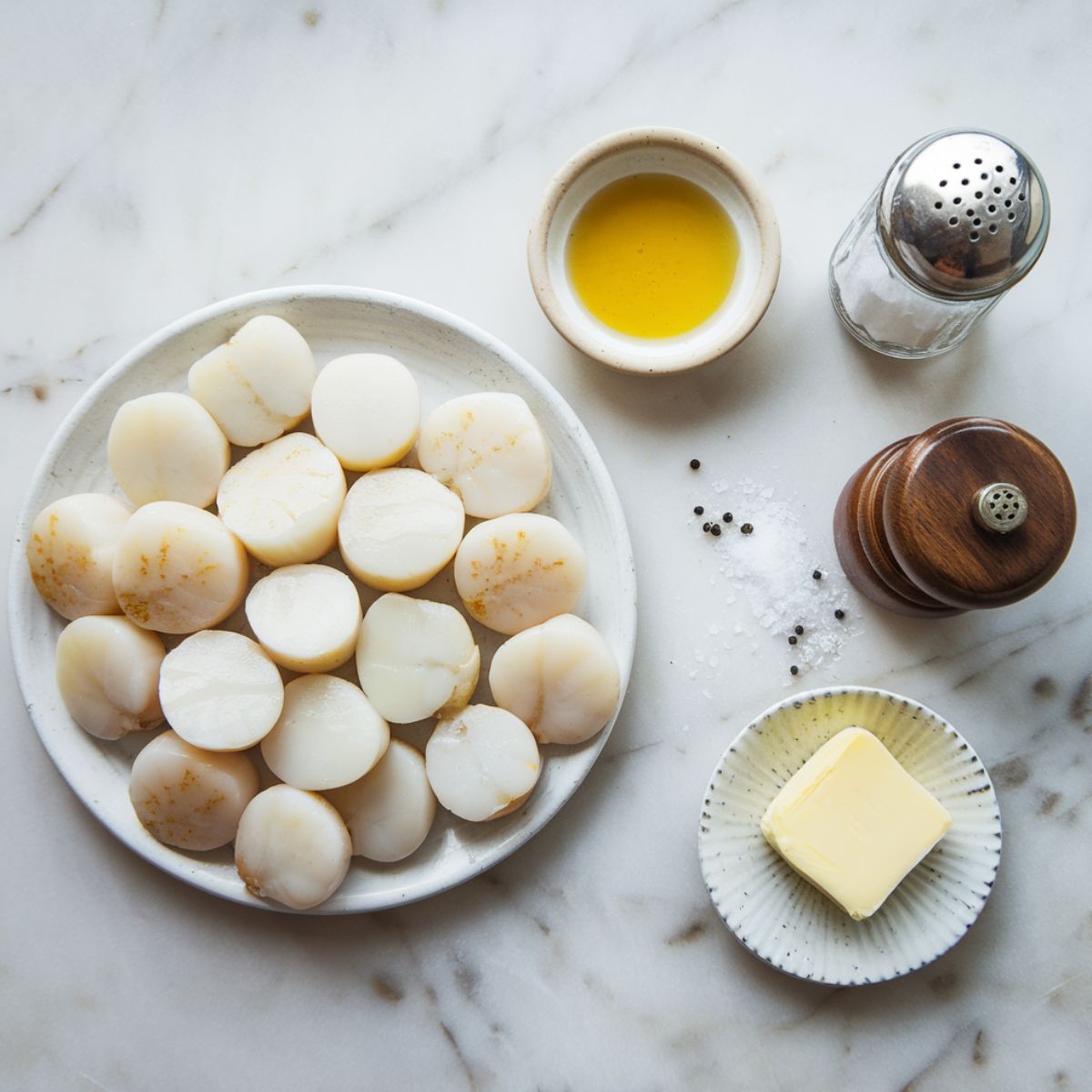 Overhead view of 12-15 fresh, white sea scallops arranged on a marble countertop, with a small dish of vegetable oil or clarified butter, a rustic salt shaker, a classic pepper grinder, and a pat of unsalted butter displayed nearby.