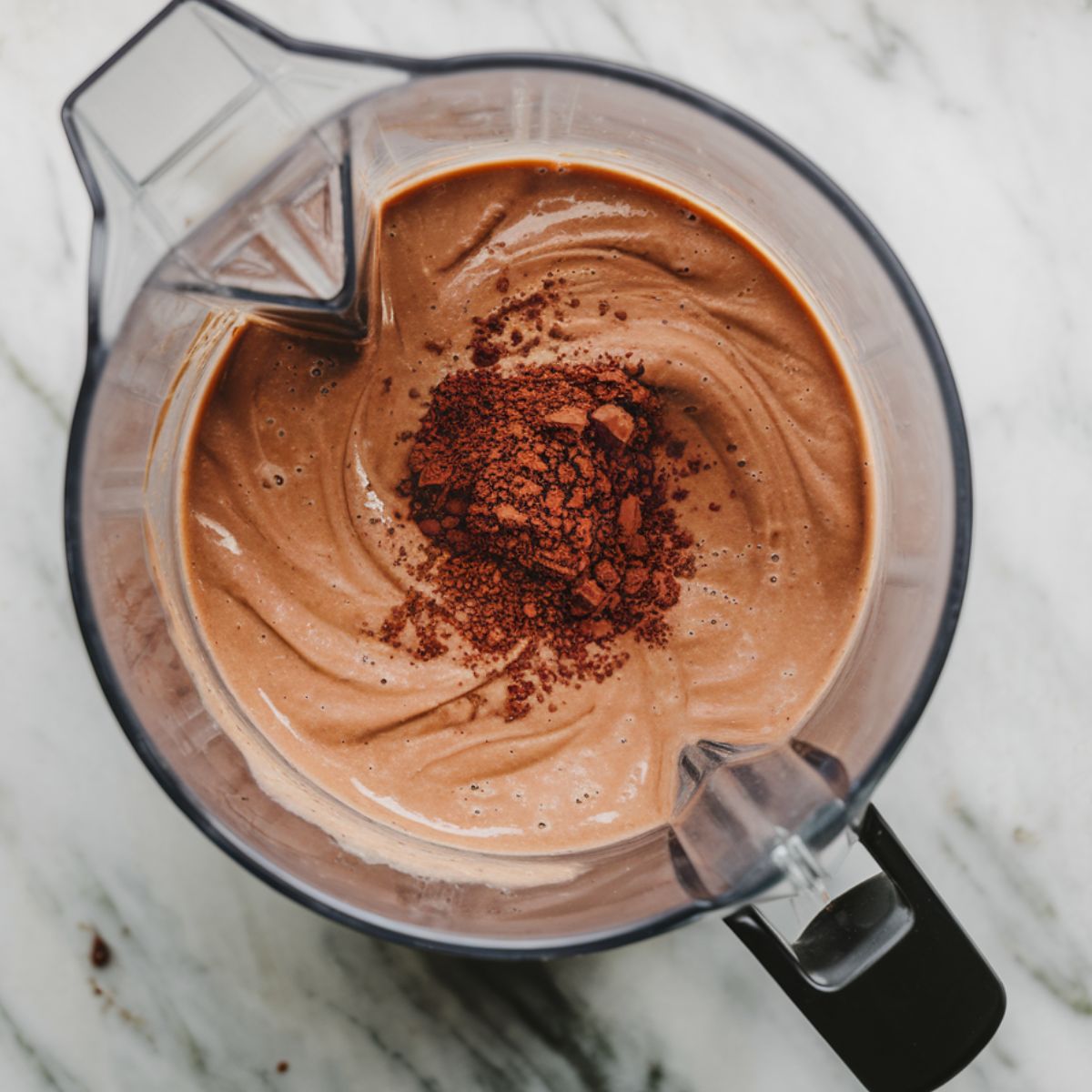 Blender on a marble countertop filled with a smooth, creamy mixture of almond milk, chocolate protein powder, peanut butter, and cocoa powder.