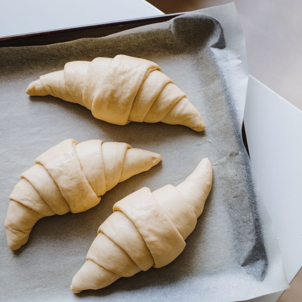 Top-down view of a white kitchen counter with a parchment-lined tray holding three freshly rolled croissant-shaped dough pieces.