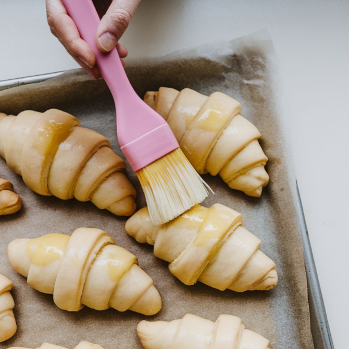 Parchment-lined baking sheet with unbaked croissants being brushed with egg wash using a pink pastry brush on a clean, neutral surface.