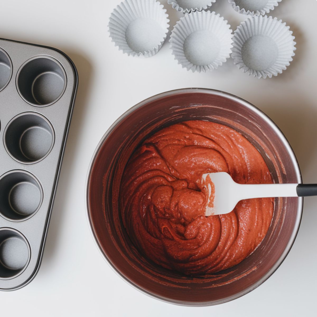 a clean white countertop, a muffin tin, white cupcake liners, a bowl of red batter, and a spatula. An oven with lighted elements is in the background.