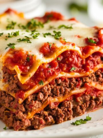 Close-up of a lasagna slice showing thick, saucy layers of ground beef and tomato sauce, gooey cheese on top, and fresh chopped parsley garnish, captured in bright lighting with a softly blurred background.