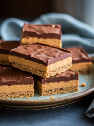 Close-up of homemade no-bake peanut butter bars with a glossy chocolate topping on a rustic ceramic plate, set against a soft blue linen napkin.