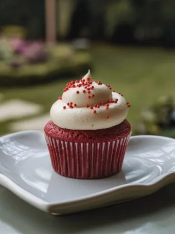 Close-up of a red velvet cupcake with white cream cheese frosting, topped with red sprinkles, placed on a heart-shaped white plate.