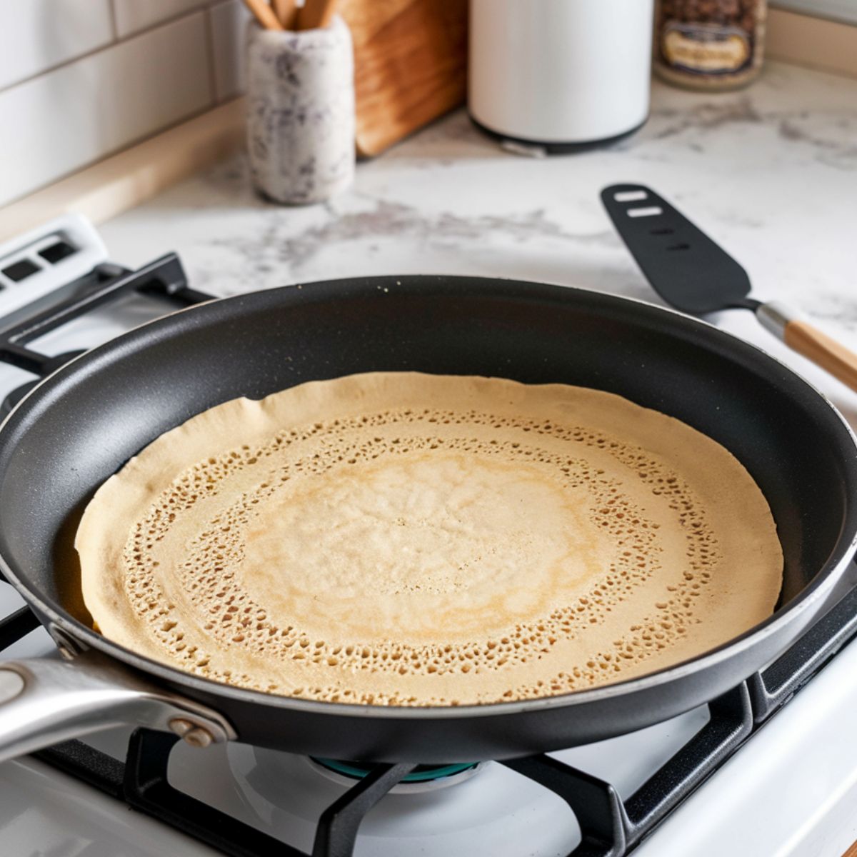 A non-stick crepe pan with a thin crepe cooking, edges slightly browning, with a spatula and batter-filled ladle nearby on a white marble countertop.