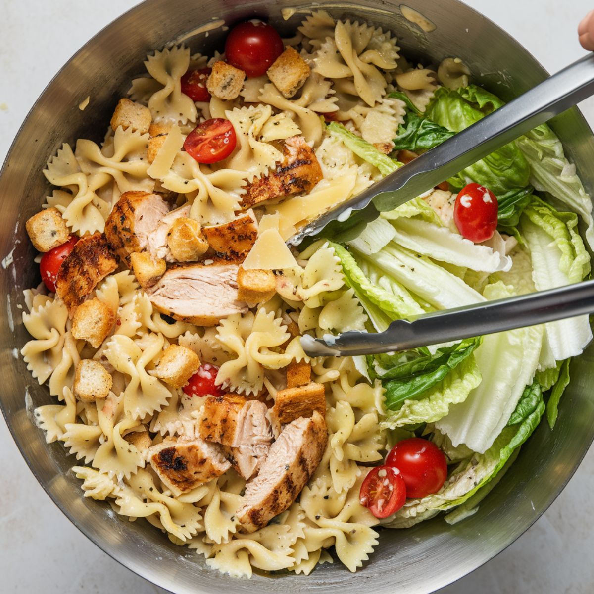 A mixing bowl with pasta, chicken, romaine lettuce, and dressing being tossed with tongs.