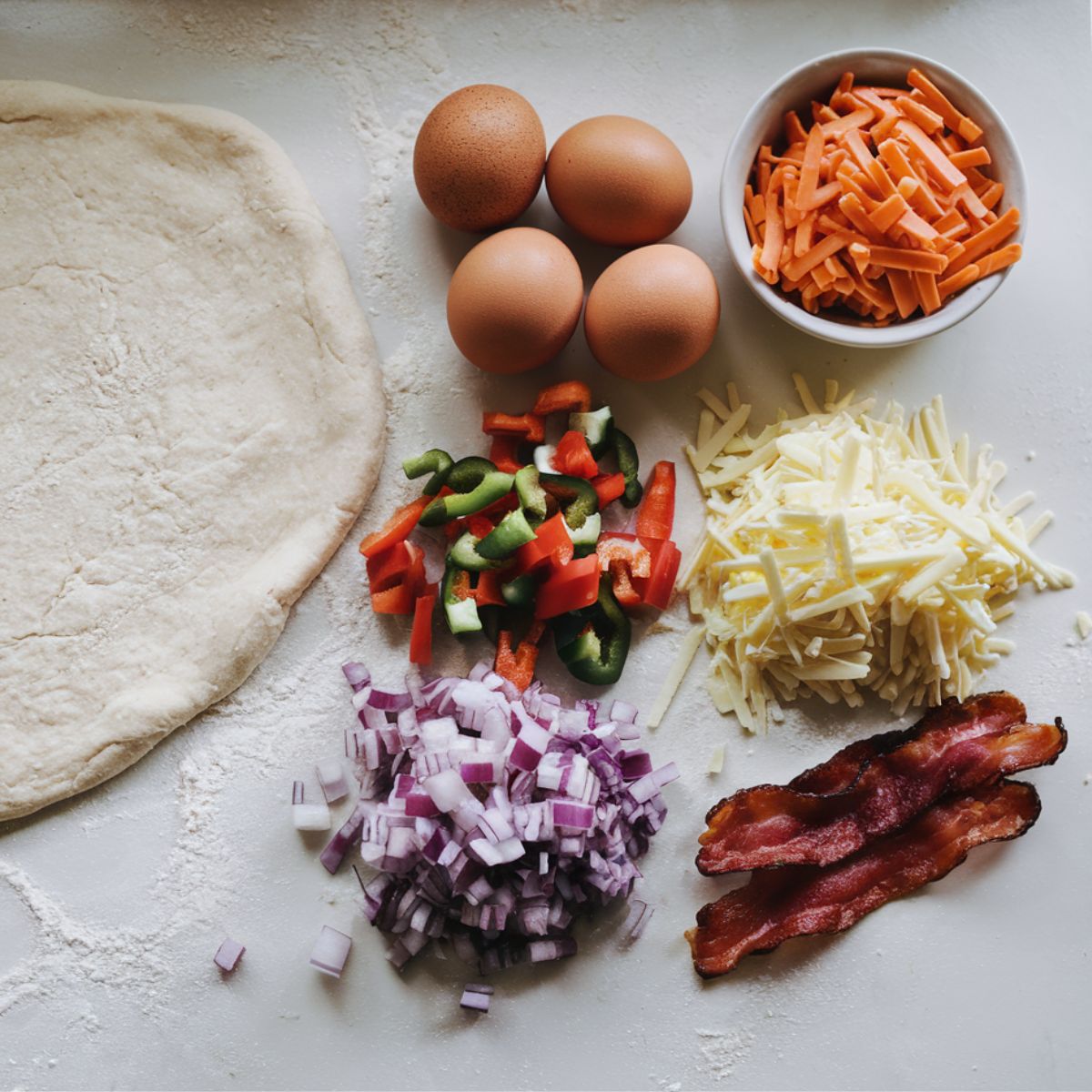  Fresh ingredients for breakfast pizza bites spread out on a white kitchen counter, including pizza dough, eggs, cheese, vegetables, and bacon.