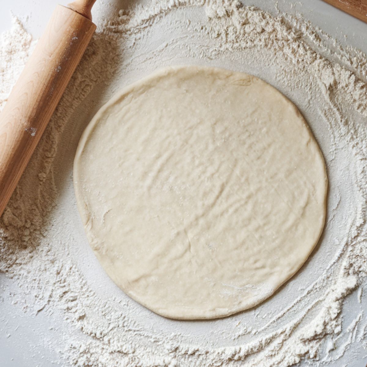 A round piece of pizza dough rolled out on a lightly floured surface, with a wooden rolling pin nearby. The dough has a slightly uneven texture, giving it a homemade feel.