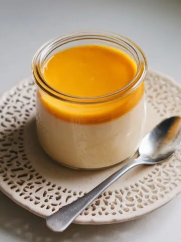 : Homemade mango panna cotta in a glass jar with a golden mango topping on a white kitchen counter.