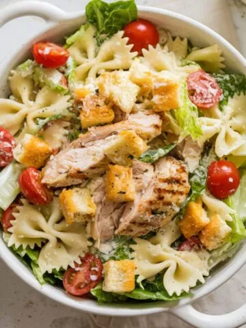 A bowl of freshly made Chicken Caesar Pasta Salad Recipe with crispy romaine, grilled chicken, cherry tomatoes, parmesan, and croutons on a white kitchen counter.