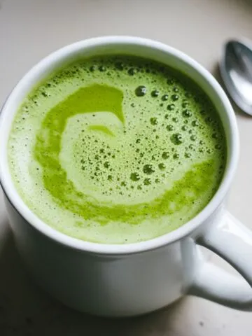 Homemade Starbucks Matcha Green Tea Latte recipe in a mug, frothy and vibrant green, with matcha powder and a whisk on a white kitchen counter.