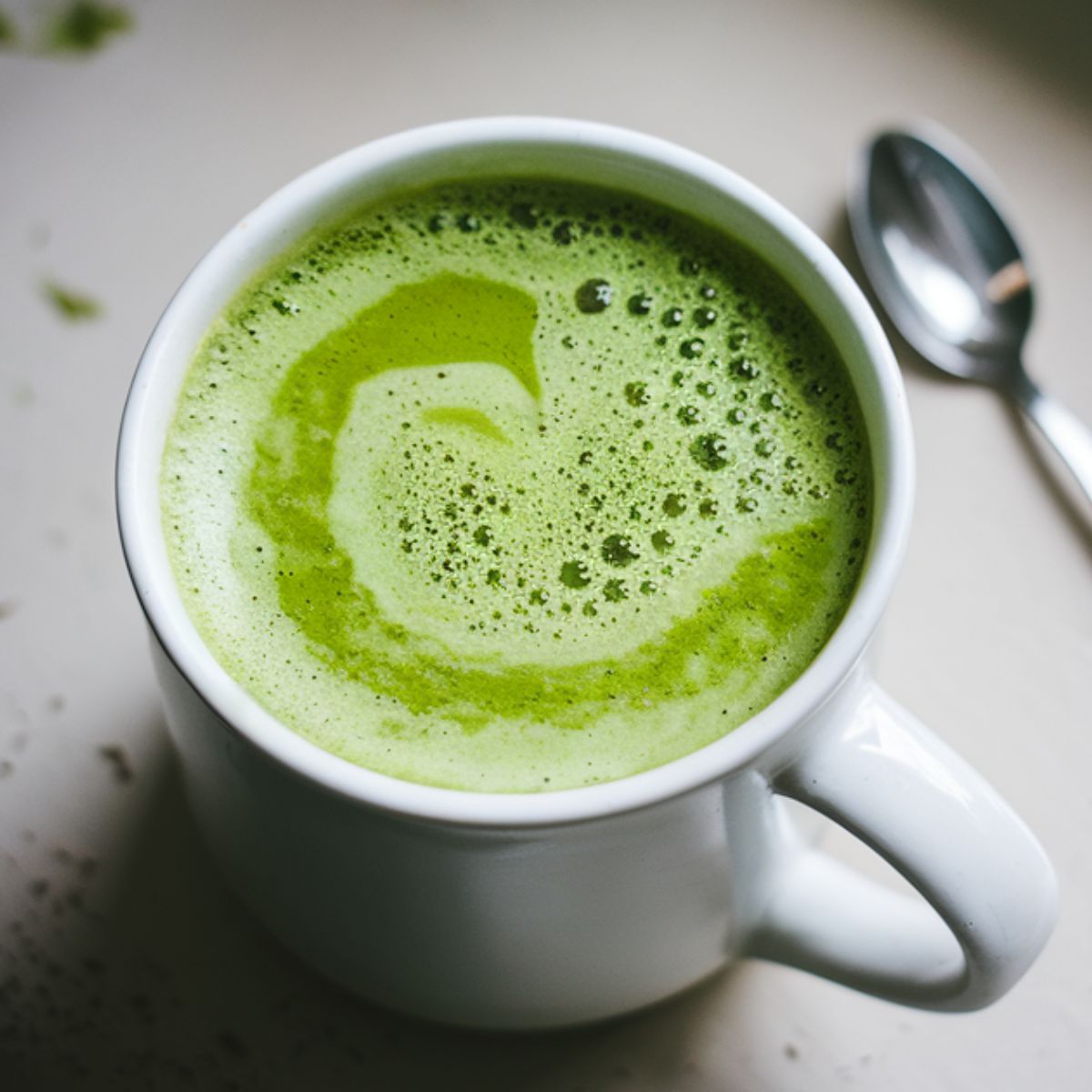 Homemade Starbucks Matcha Green Tea Latte recipe in a mug, frothy and vibrant green, with matcha powder and a whisk on a white kitchen counter.