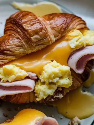 A golden, flaky croissant sandwich filled with ham, melted cheese, and spinach, served on a plate.