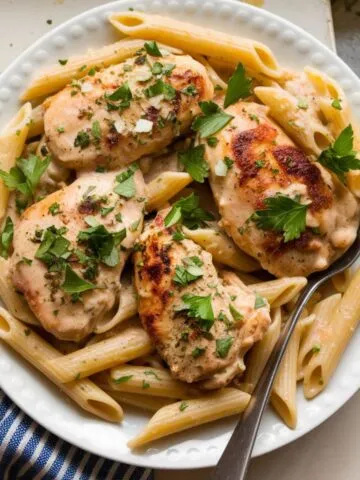 A bowl of creamy garlic parmesan chicken pasta with golden-brown chicken pieces and a creamy white sauce, garnished with fresh parsley, served on a white plate on a home kitchen counter.