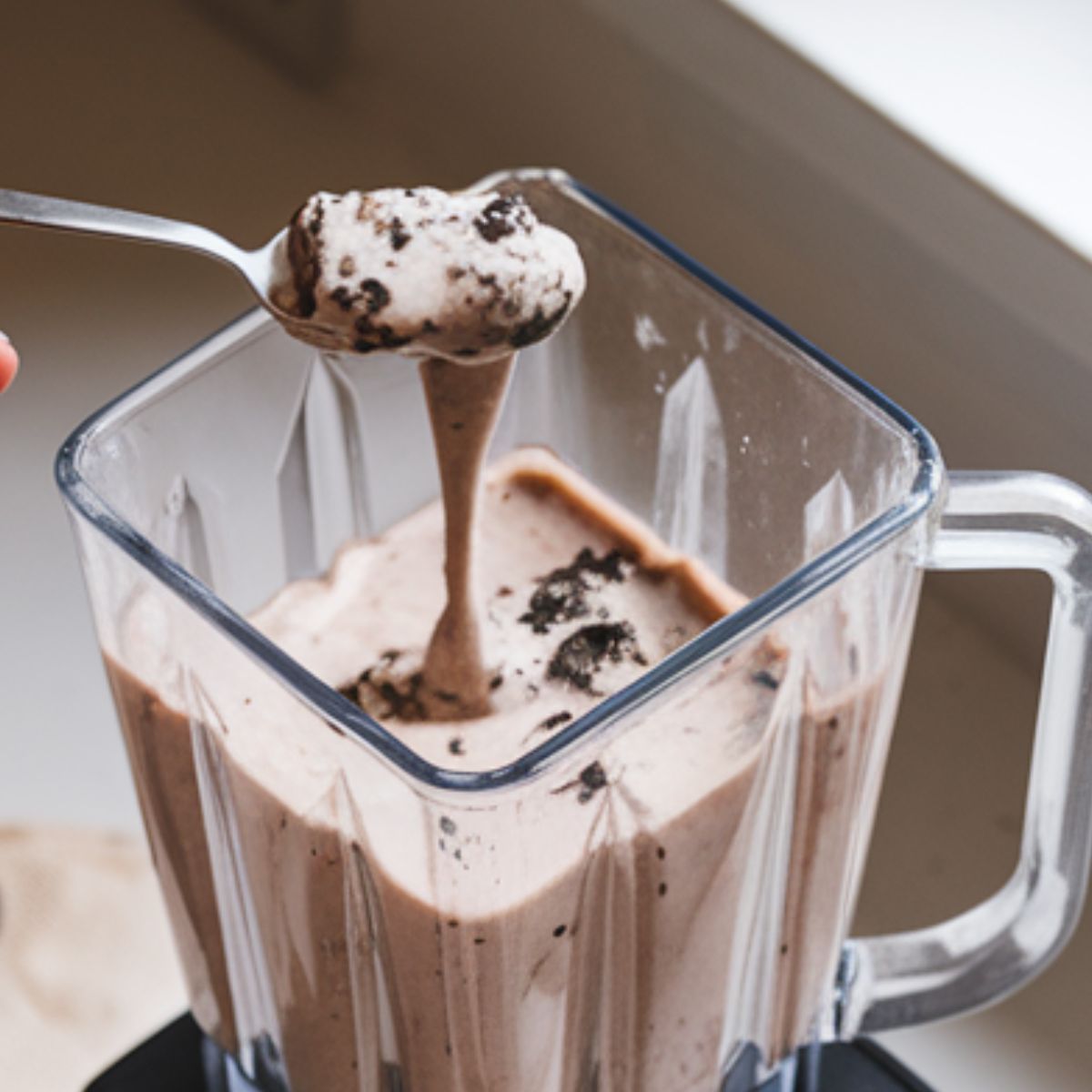 A spoon lifting thick Oreo milkshake from a blender to check consistency.