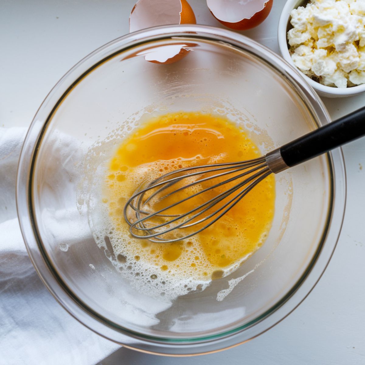  A bowl of whisked eggs with a fork, a bowl of cottage cheese, and chopped chives on a white kitchen counter, with eggshells nearby for a homemade touch.