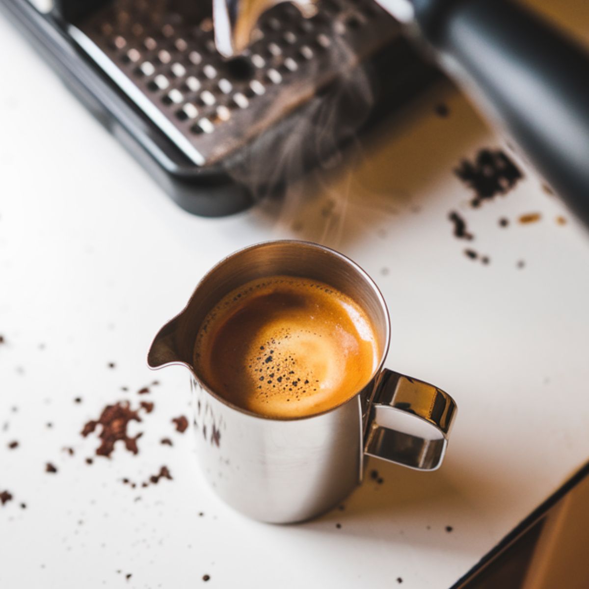 A stainless steel espresso-pouring cup filled with fresh espresso, topped with golden crema, on a white kitchen counter with scattered coffee grounds.