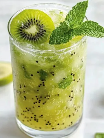 A tall glass of kiwi mint mojito garnished with mint and kiwi slices, sitting on a white kitchen counter.