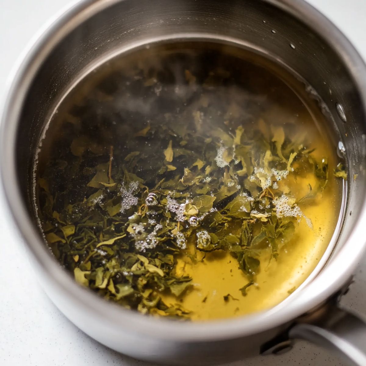  Loose Green tea steeping in a small saucepan on a white counter with steam rising gently.