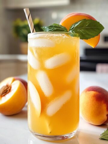 A homemade glass of peach green tea lemonade with fresh peach and lemon garnish on a white countertop.