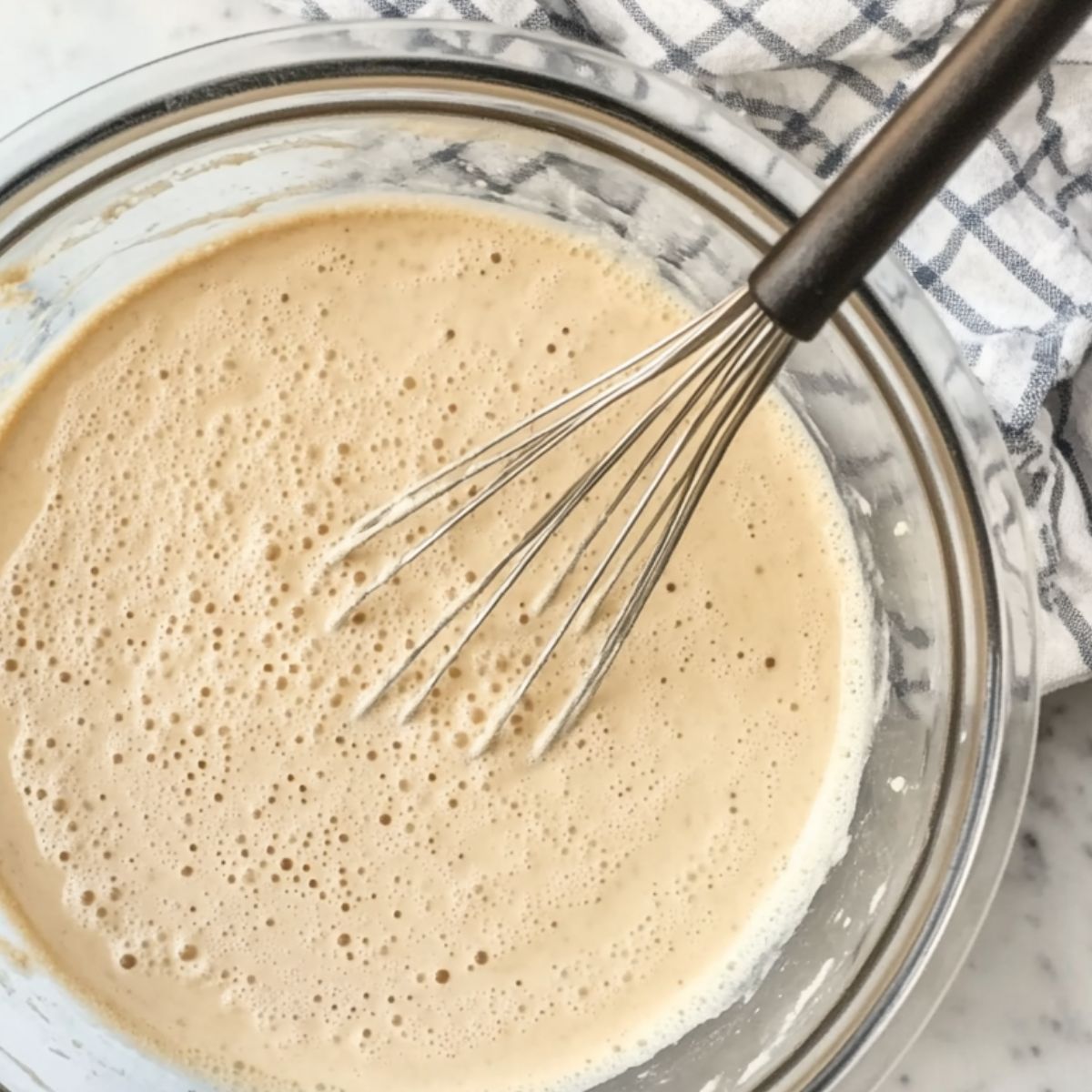 Bowl with crepe batter being whisked on a white kitchen counter
