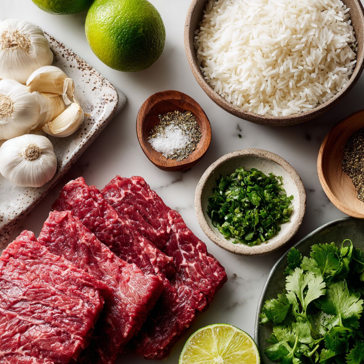  Raw steak, lime, garlic, fresh cilantro, spices, and rice on a white counter before cooking
