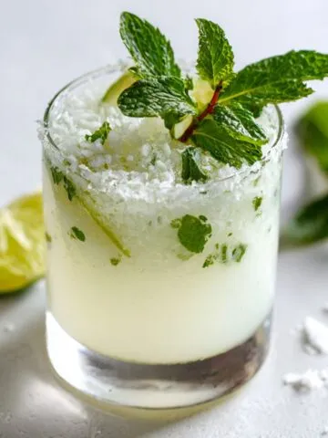 Coconut mojito recipe in a highball glass with crushed ice, lime wedge and mint sprig on a white kitchen counter.