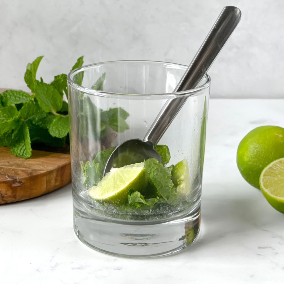 Mint and lime being muddled in a glass on a white kitchen counter.