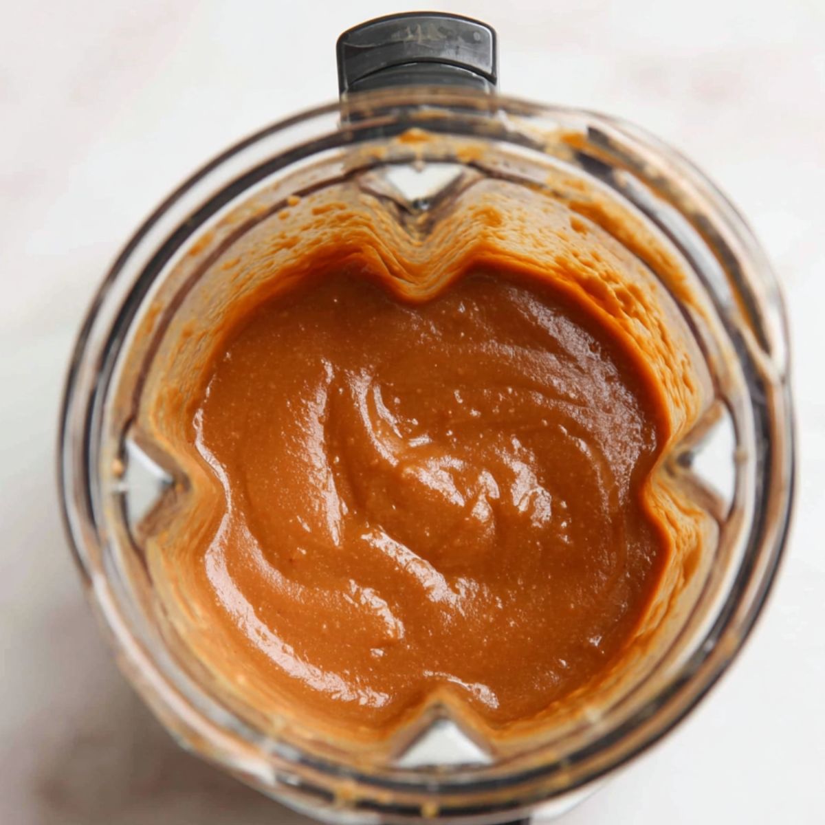Blender filled with creamy date mixture as almond butter, vanilla, and sea salt . on a white kitchen counter