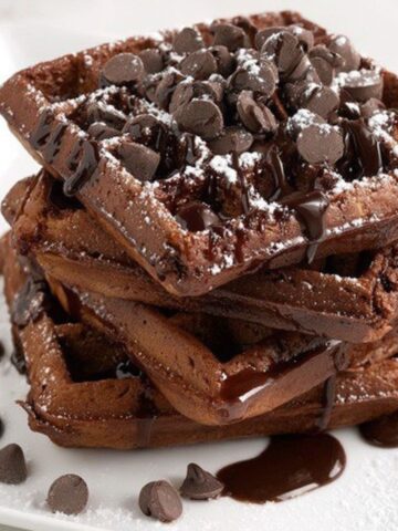 A stack of imperfectly shaped homemade brownie waffles with Chocolate sauce on a white kitchen counter.