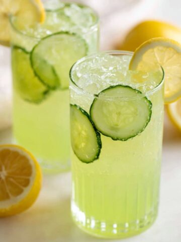 Cups of cucumber lemonade on a white kitchen counter with fresh lemon and cucumber slices