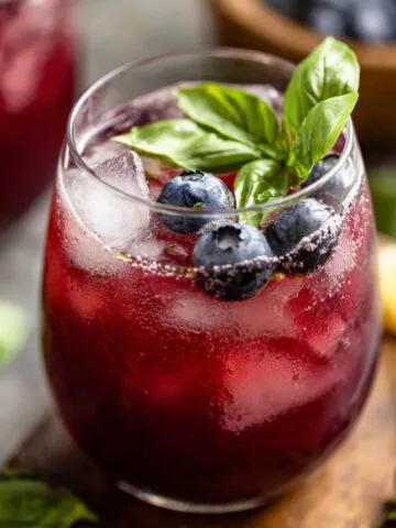 A homemade glasses of blueberry basil lemonade with fresh blueberries, basil leaves, and ice on a wooden kitchen counter