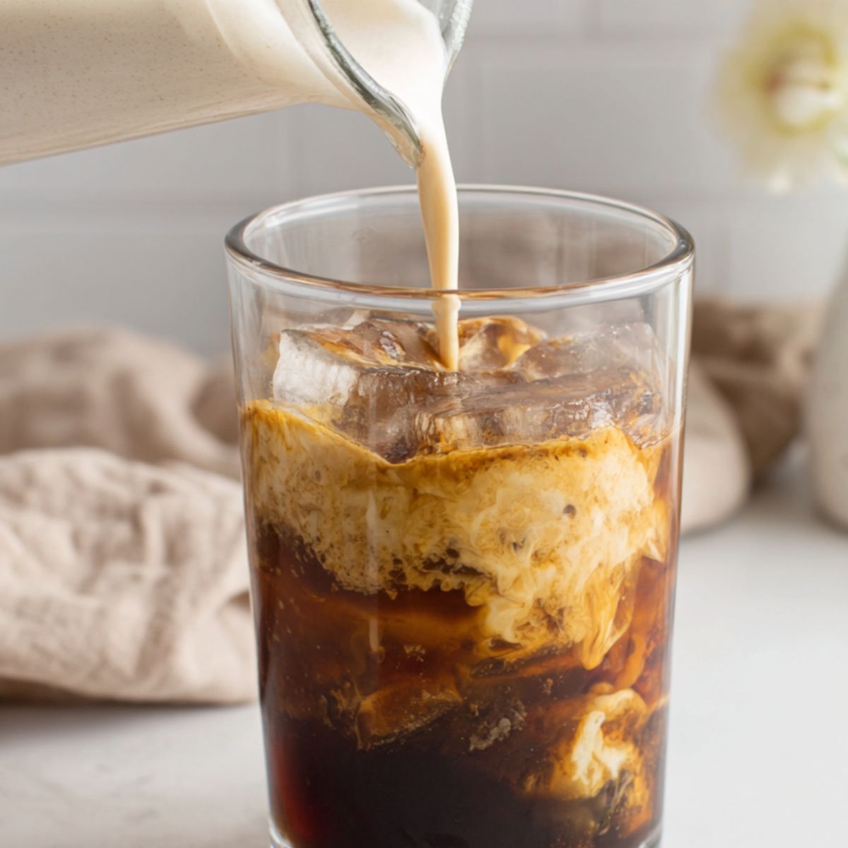 Oat milk being poured into a glass of iced espresso on a kitchen counter.