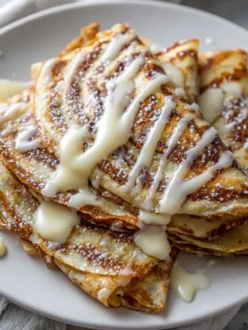 Freshly stacked cinnamon roll crepes with glaze drizzle on white plate