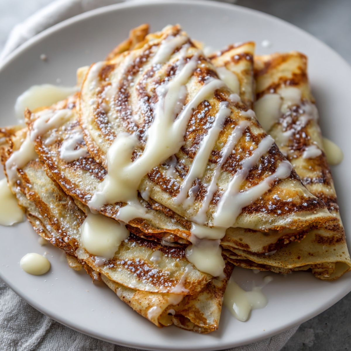Freshly stacked cinnamon roll crepes with glaze drizzle on white plate