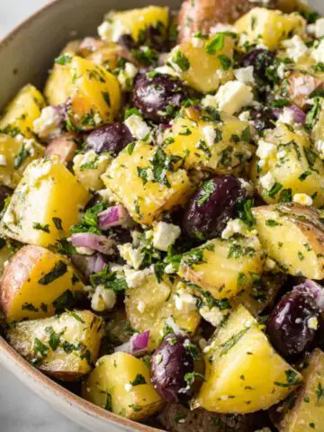 Homemade Greek potato salad with feta, olives, and herbs served in a bowl on a white kitchen counter.
