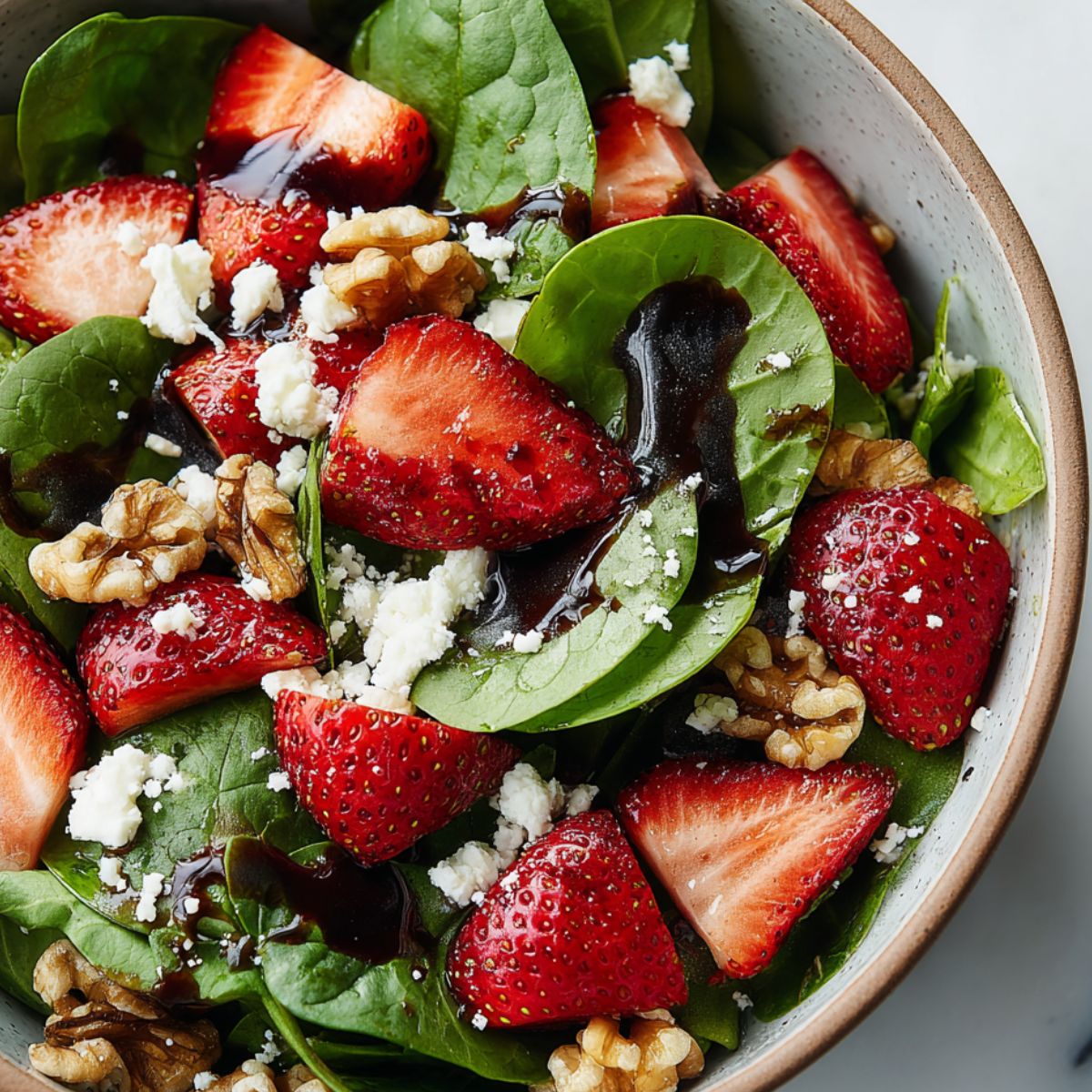 Overhead view of homemade walnut strawberry spinach salad in a ceramic bowl on a white kitchen counter.