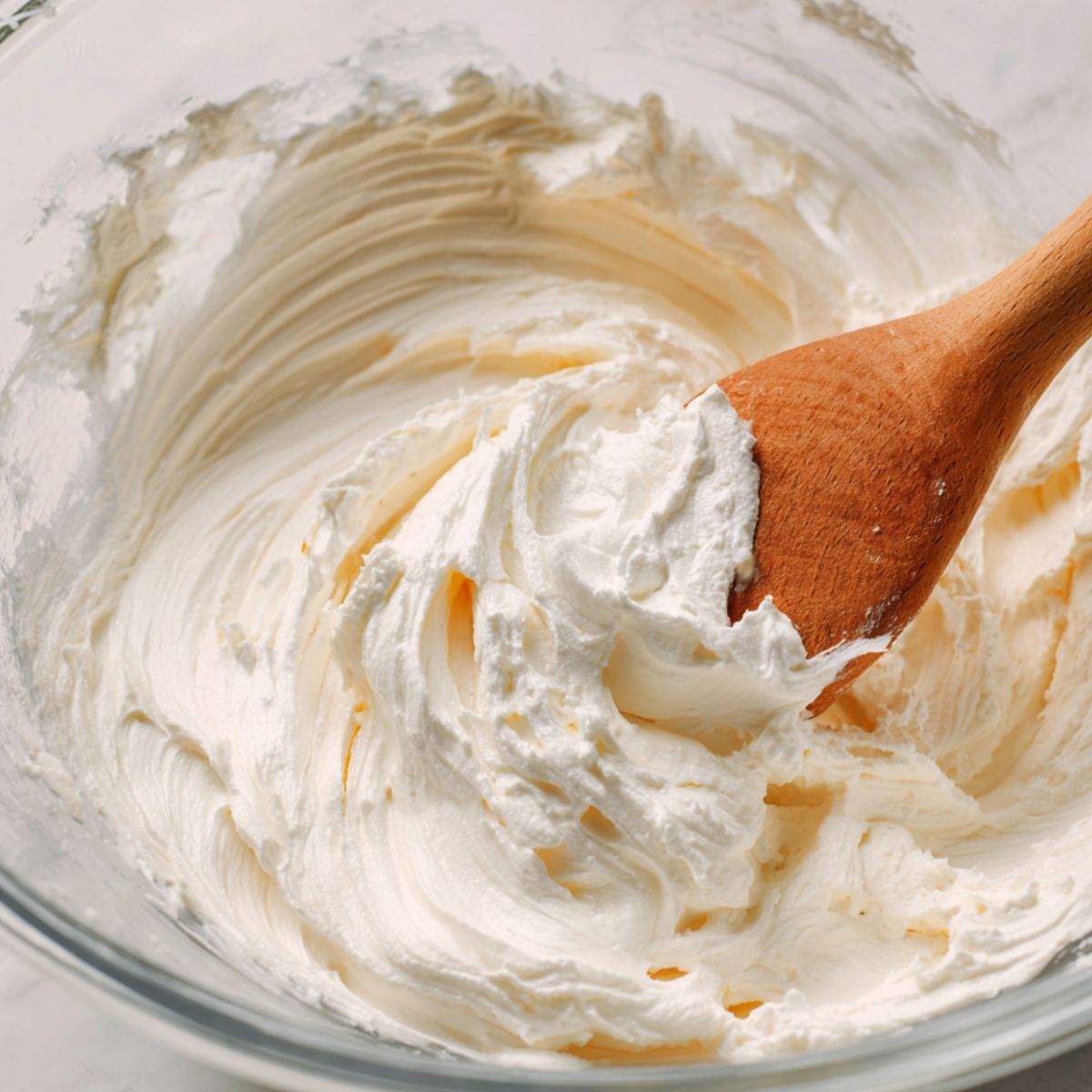Folding whipped cream into cream cheese mixture in a glass bowl, top-down view.